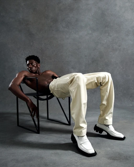 A shirtless Black male fashion model poses sitting in a chair wearing glossy off-white pants and white penny loafers against a grey backdrop.
