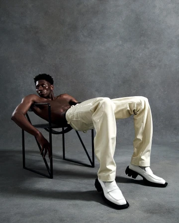 A shirtless Black male fashion model poses sitting in a chair wearing glossy off-white pants and white penny loafers against a grey backdrop.
