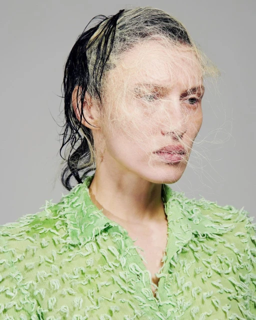 Woman's portrait with sheer green shirt and facial covering of white fiber strands against a plain grey background in a studio shot.
