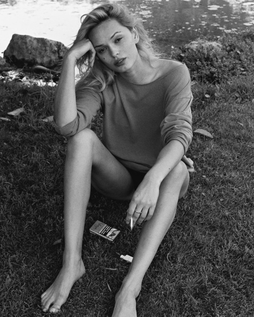Black and white shot of a blonde woman sitting on grass by a body of water, holding a cigarette, with a cigarette pack nearby.