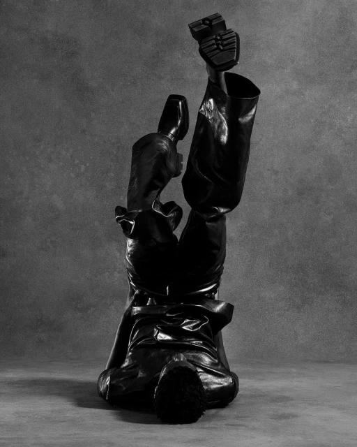 Monochrome fashion shot of a figure in shiny black trousers and boots doing a handstand against a mottled grey background.