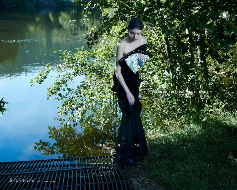 A woman in a dark dress stands near a river surrounded by lush greenery, creating a serene fashion shot in a natural setting.