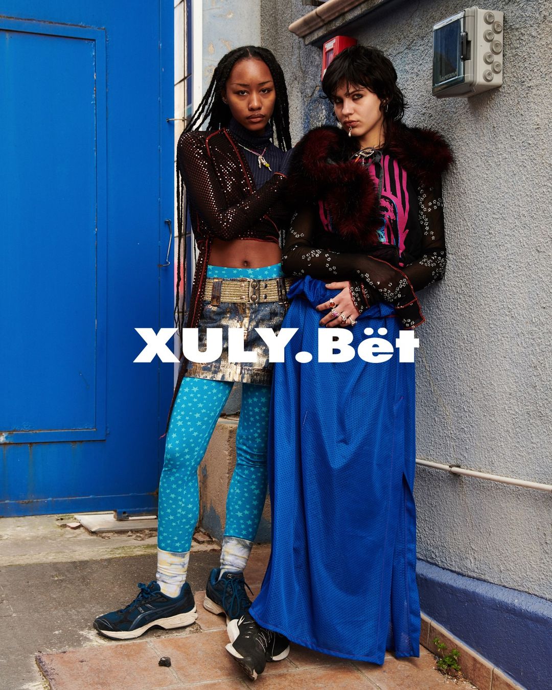 Two stylish adult women pose outdoors in fashionable clothing featuring faux fur, unique leggings, skirts, and sneakers in an urban setting.
