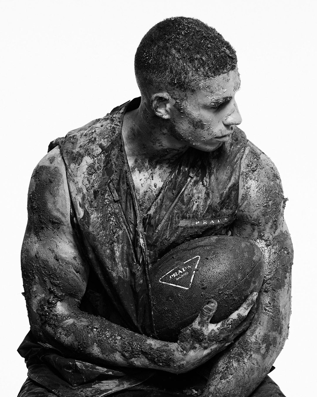 Gritty portrait of a man covered in mud holding a football with a Prada logo against a stark white background in black and white.