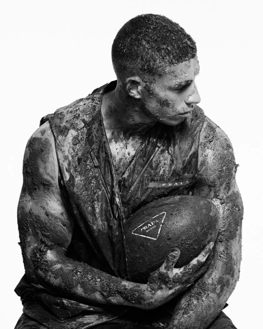 Gritty portrait of a man covered in mud holding a football with a Prada logo against a stark white background in black and white.