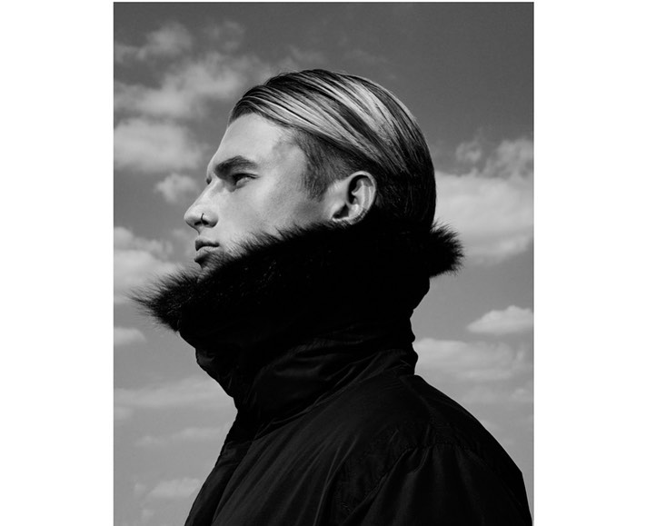 Monochrome portrait of an adult male with blond highlighted hair, wearing a dark winter coat with a fur collar, against a cloudy sky.