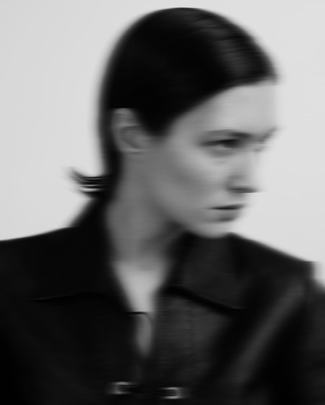 Monochrome portrait of a woman with slicked-back dark hair wearing a dark jacket against a neutral background with blurred focus.