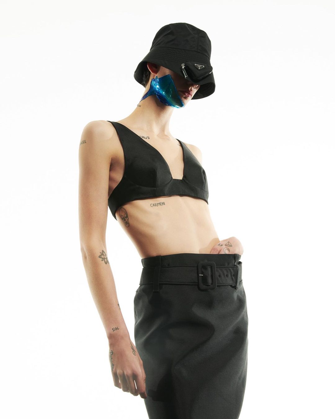 Stylish fashion portrait of a slim woman in black bra top, bucket hat, and skirt against a bright background in a studio setting.