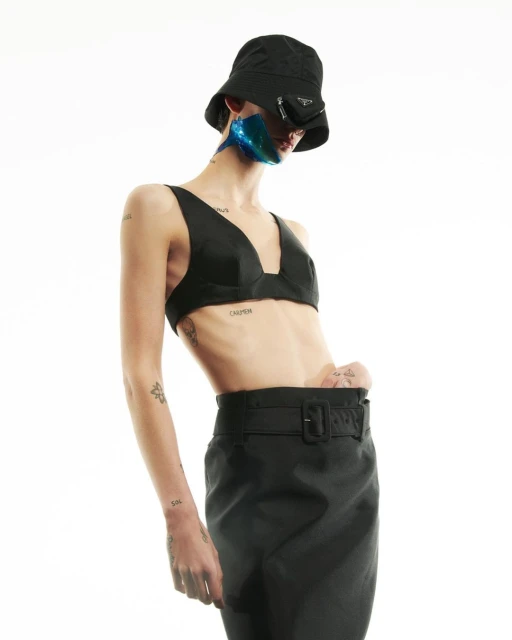 Stylish fashion portrait of a slim woman in black bra top, bucket hat, and skirt against a bright background in a studio setting.