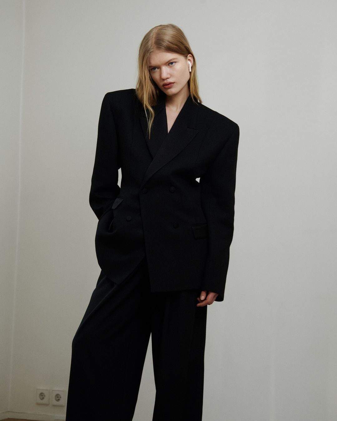 Blonde woman in black blazer and wide-leg pants posing against a plain white wall, showcasing a modern and minimalist fashion aesthetic.