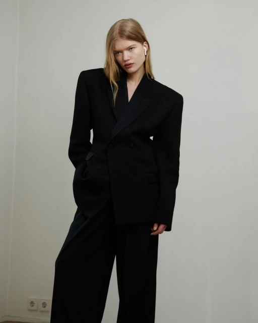 Blonde woman in black blazer and wide-leg pants posing against a plain white wall, showcasing a modern and minimalist fashion aesthetic.