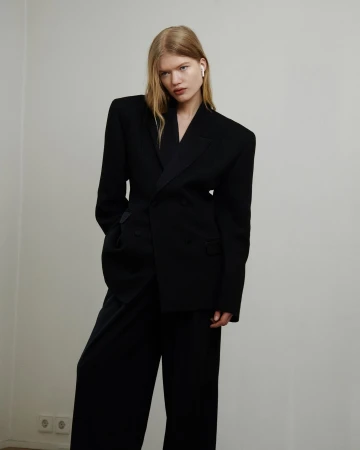 Blonde woman in black blazer and wide-leg pants posing against a plain white wall, showcasing a modern and minimalist fashion aesthetic.