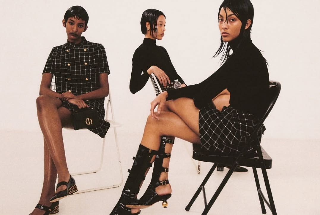 Three women in patterned dresses and stylish shoes pose against a white backdrop, seated on black and white chairs, for a fashion editorial.