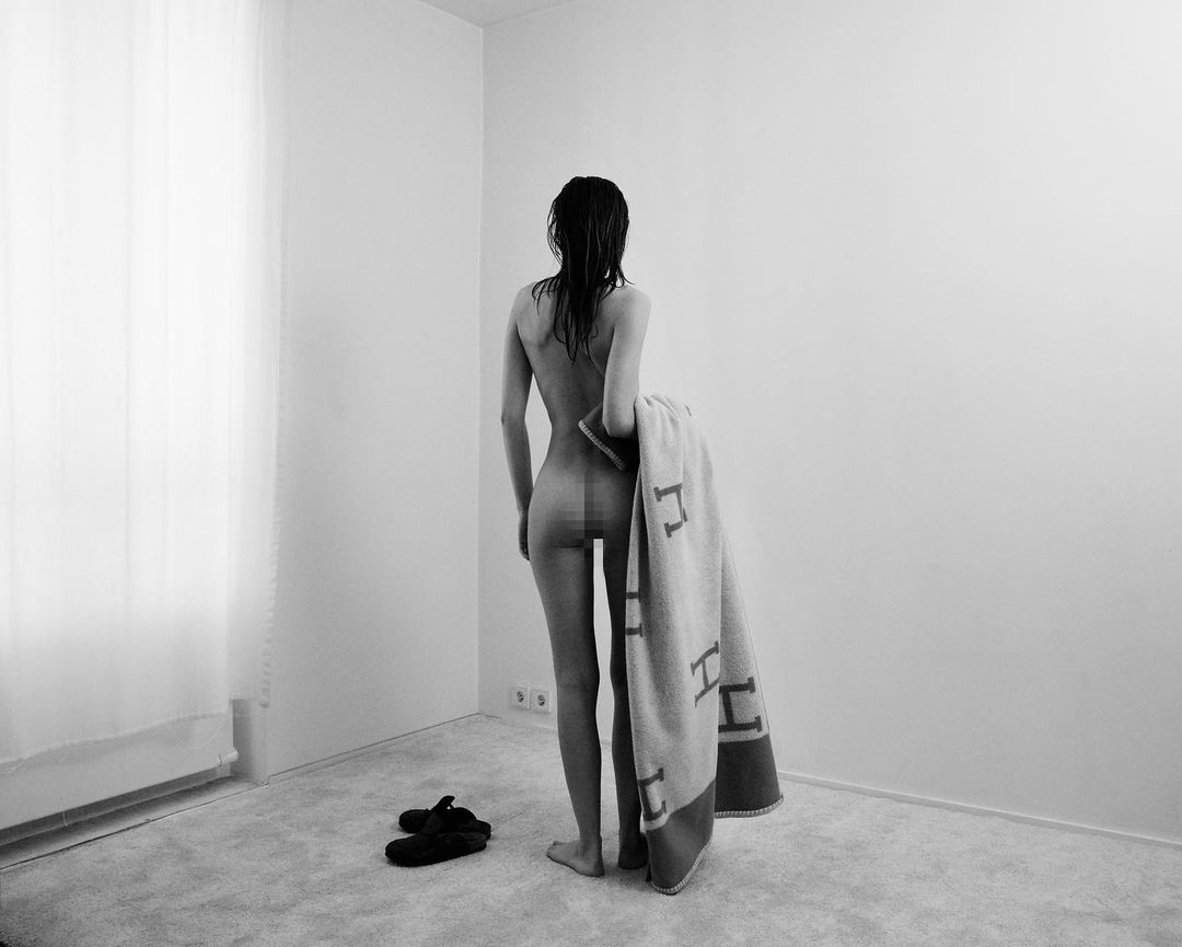 Black-and-white indoor shot of a partially nude woman with wet hair holding a towel, standing near a pair of shoes and a window.
