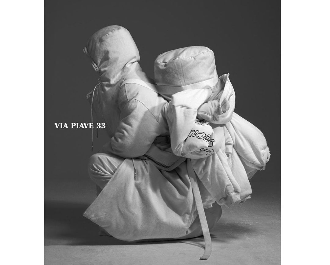 Monochrome image of two people in hooded coats, one kneeling with the other leaning on them, showcasing fashion and style in studio.