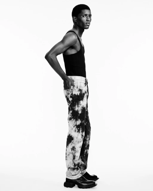 Fashionable adult male model poses in black tank top, tie-dye jeans, and black leather boots in a full shot against a white backdrop.