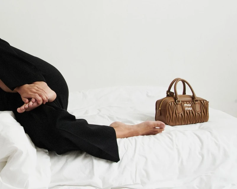 A brown leather Miu Miu handbag sits on a white bed next to a woman in black pants with bare feet. Minimalist fashion photography.