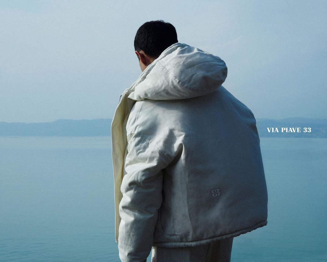 Back view of person in off-white hooded jacket overlooking blue lake and distant mountains on a bright, sunny day.