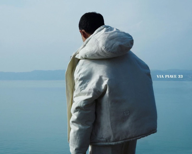 Back view of person in off-white hooded jacket overlooking blue lake and distant mountains on a bright, sunny day.