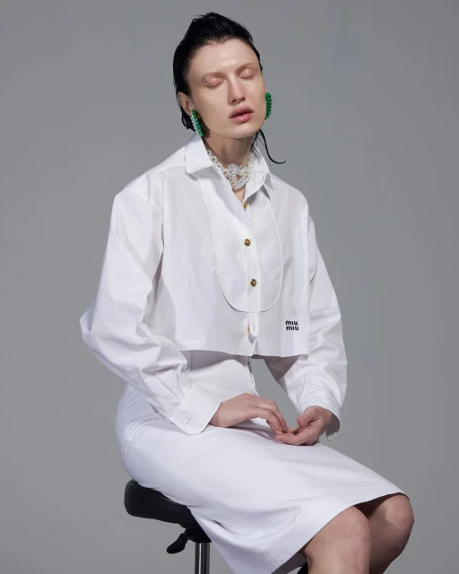 Woman with wet hair sits on a stool in a photo studio, wearing a white cropped shirt and skirt with statement earrings and necklace.