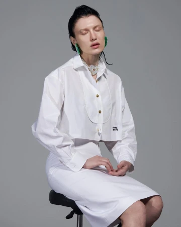 Woman with wet hair sits on a stool in a photo studio, wearing a white cropped shirt and skirt with statement earrings and necklace.