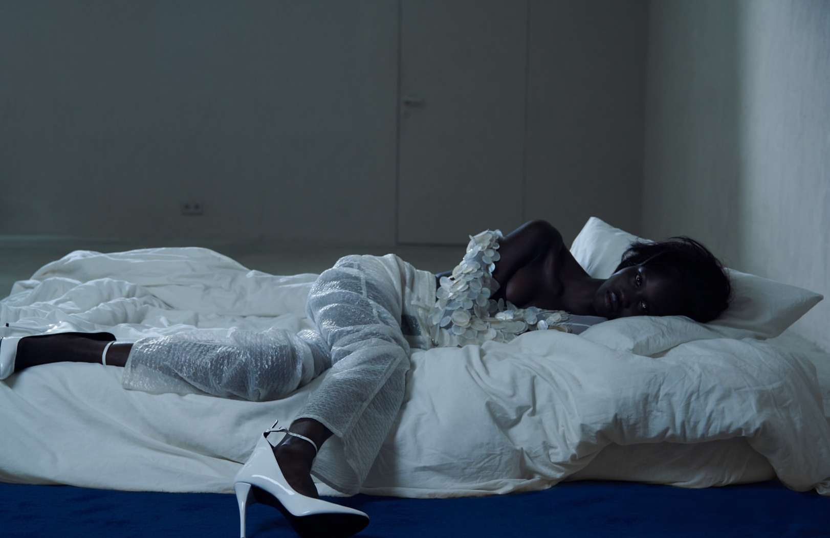 An African-American woman in an embellished top and pants with stiletto heels lies on a bed covered in white sheets in an indoor setting.