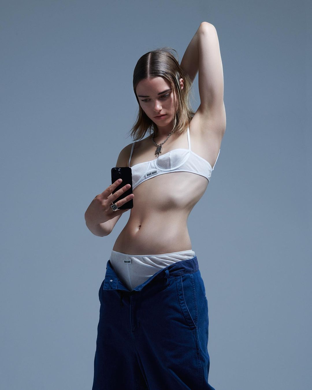 Young woman in underwear and unbuttoned jeans poses in a photo studio, holding a phone to take a selfie in front of a grey backdrop.