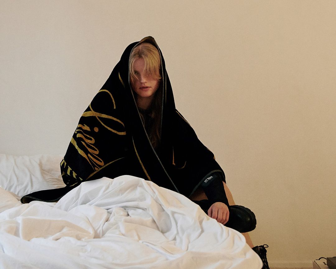 Woman sitting on a bed, wrapped in a black blanket with gold lettering, looking directly at the camera in an indoor setting.