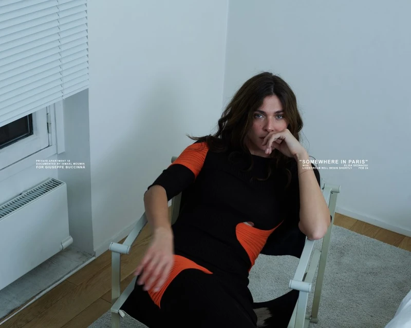 Adult woman in a black and orange dress sits on a white metal chair indoors, with light from the window and visible text.