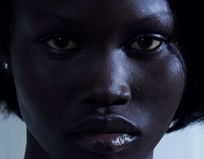 Close-up portrait of a woman with dark skin and glossy lips, looking directly at the camera, showcasing her dark hair and delicate earring.