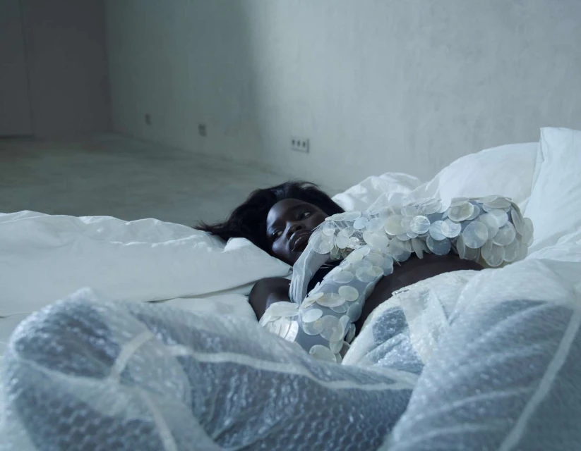 Woman lying on a bed wearing a dress covered in white discs with a clear plastic sheet over her and a concrete wall background.