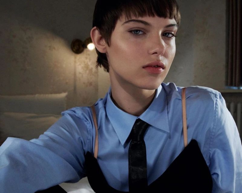 Close-up of a fair-skinned woman with a short dark hairstyle wearing a collared shirt and tie looking directly at the camera indoors.