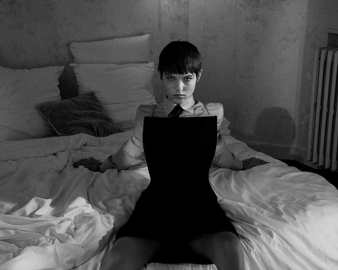 Young woman in a black dress and tie sits on a white bed with floral wallpaper in a black and white fashion photograph.