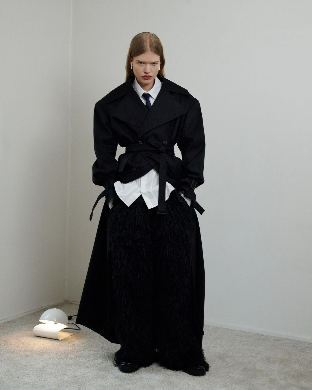 A person poses in a black overcoat with an open white shirt and a tie in a bright photo studio, standing next to a modern lamp.