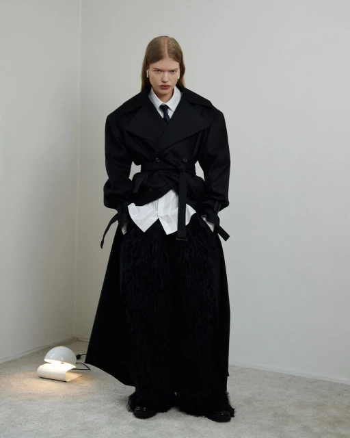 A person poses in a black overcoat with an open white shirt and a tie in a bright photo studio, standing next to a modern lamp.