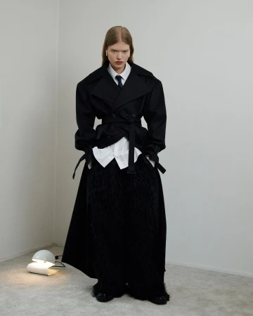 A person poses in a black overcoat with an open white shirt and a tie in a bright photo studio, standing next to a modern lamp.