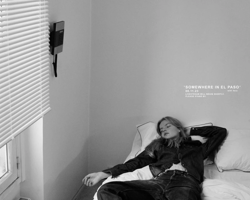 A young woman in denim lies on a bed with a white pillow against a wall with a retro phone and poster. Black and white.