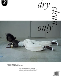 Black model Chudier Billy lying flat on a concrete floor wearing white top and trousers with white high heels, photographed by Ismael Moumin.