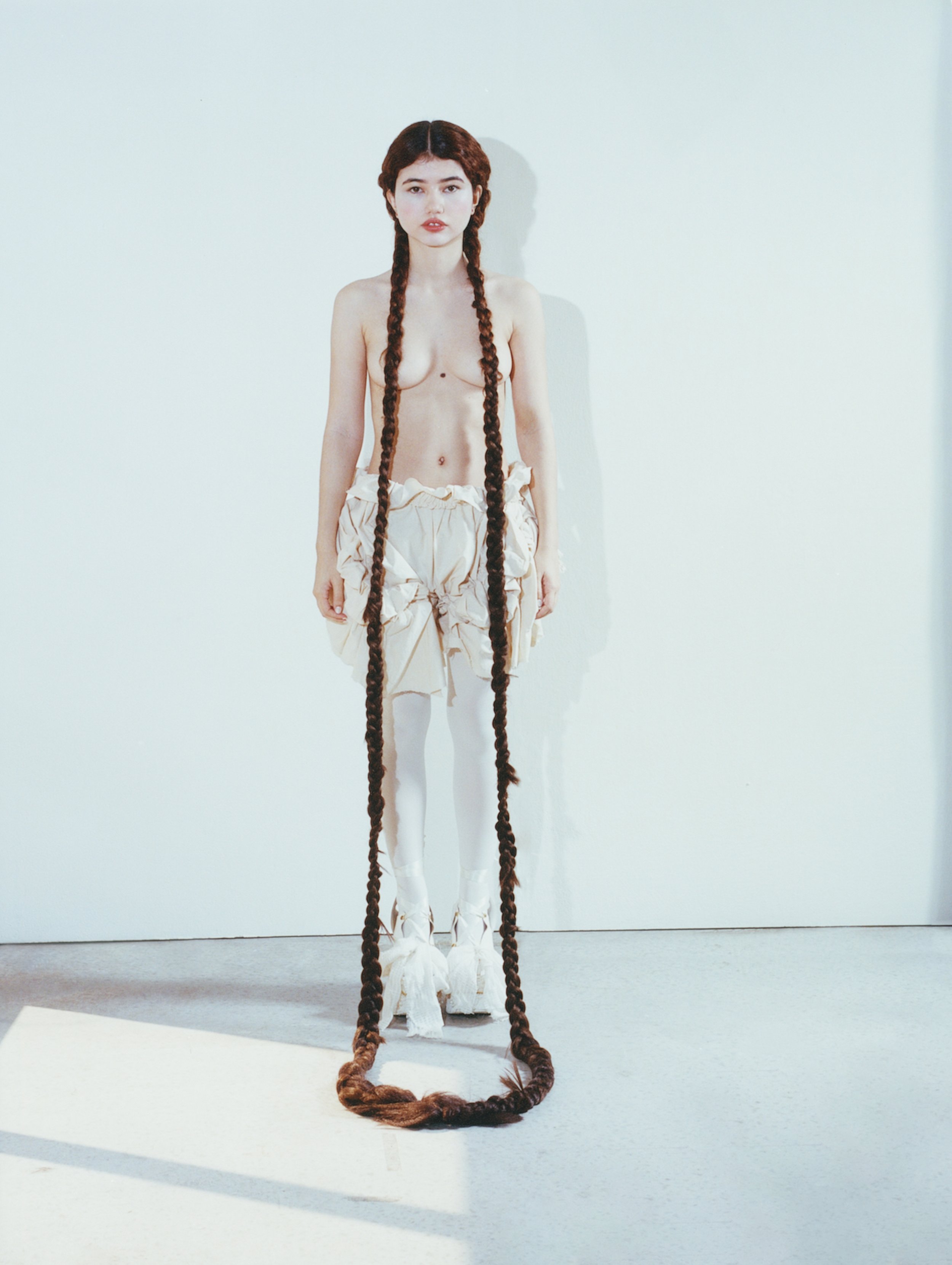 Young woman stands tall in a light studio, showcasing long braided hair, wearing linen shorts, white stockings, and shoes, bathed in soft light.