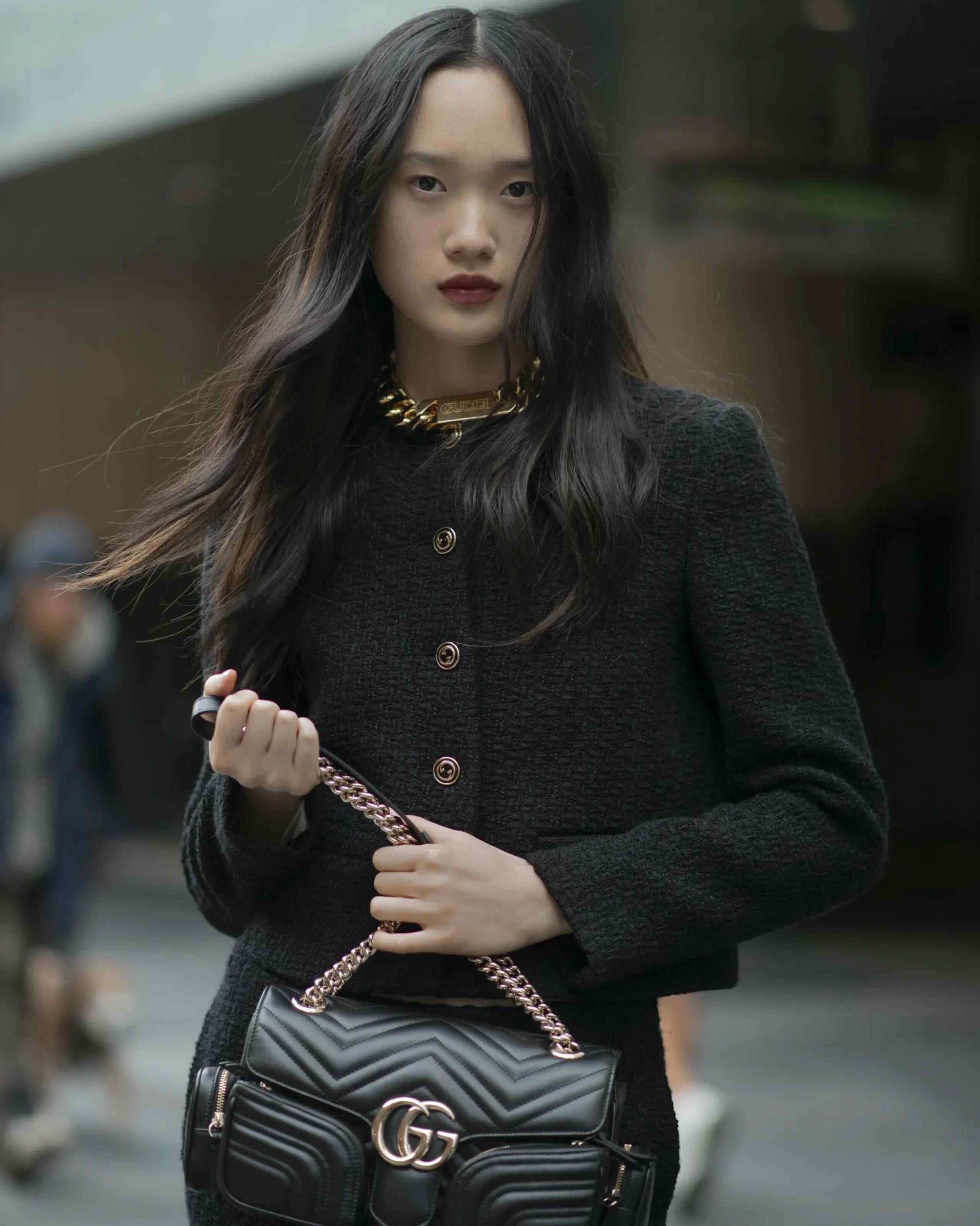 Asian woman in a dark jacket and skirt holds a black Gucci purse with a gold chain outdoors in an urban setting, medium shot.