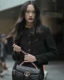 East Asian woman in black tweed jacket holding quilted leather handbag with gold chain strap outdoors