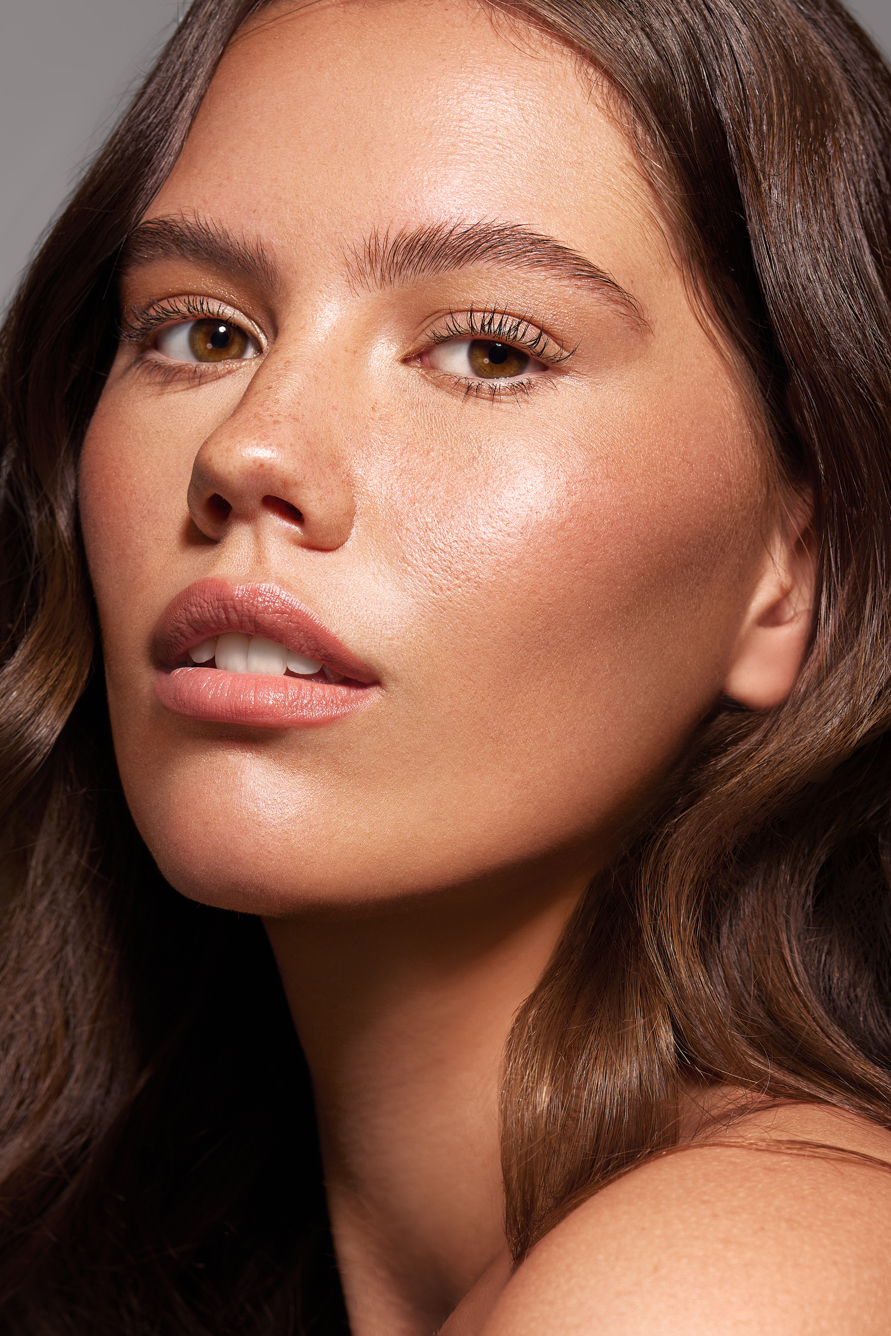 Close-up portrait of a brunette woman with fair skin and natural makeup looking at the camera against a neutral grey backdrop.