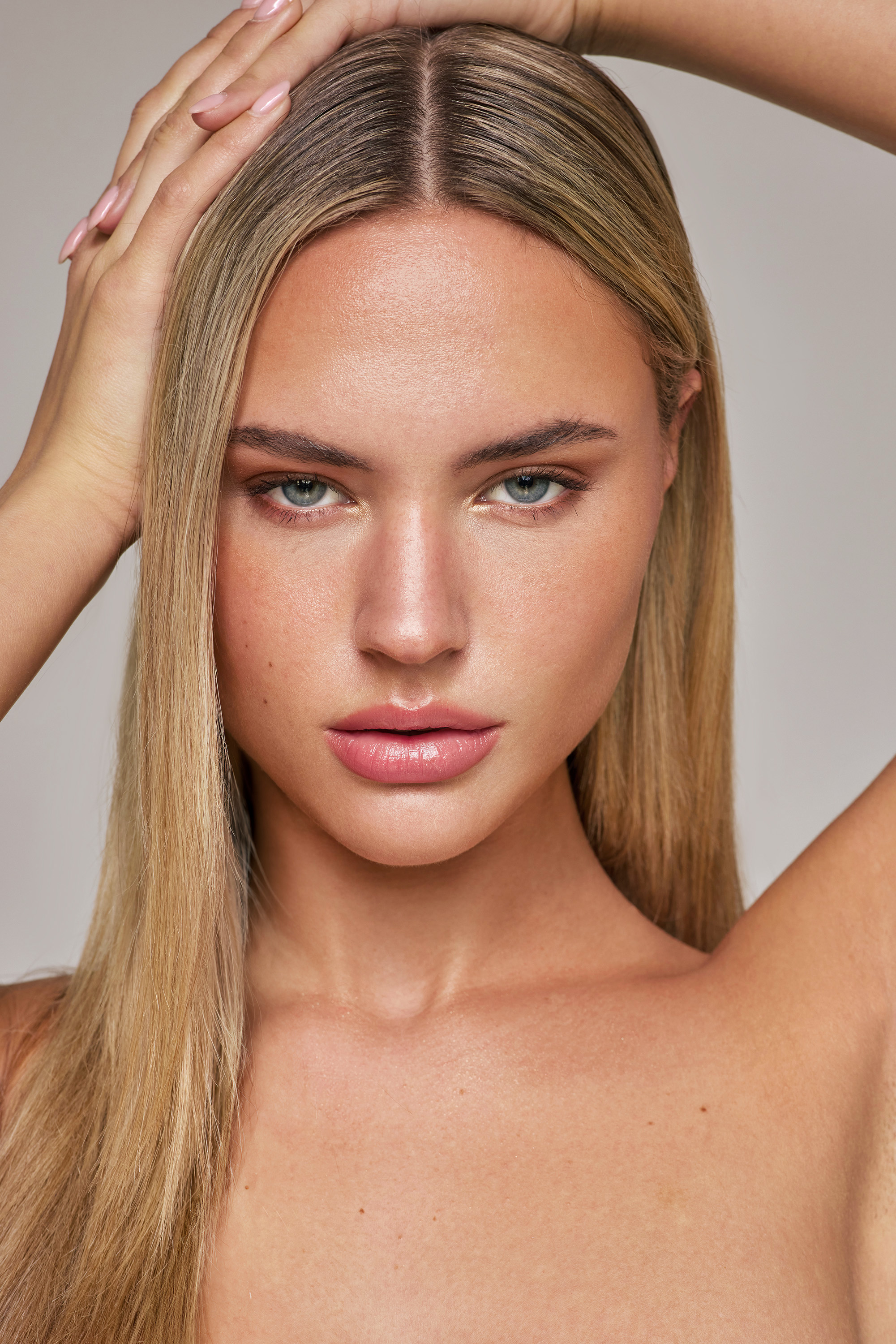 Close-up portrait of a fair-skinned adult woman with blonde hair, light eye makeup, and hands on her head, against a light neutral background.