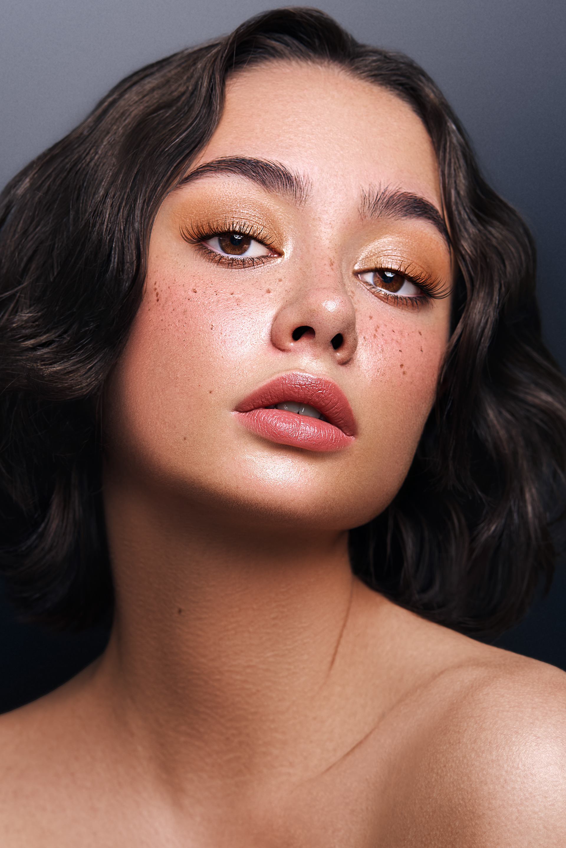 Close-up portrait of a young adult female model with dark wavy hair, wearing golden eyeshadow and nude lipstick, posing against a dark background.
