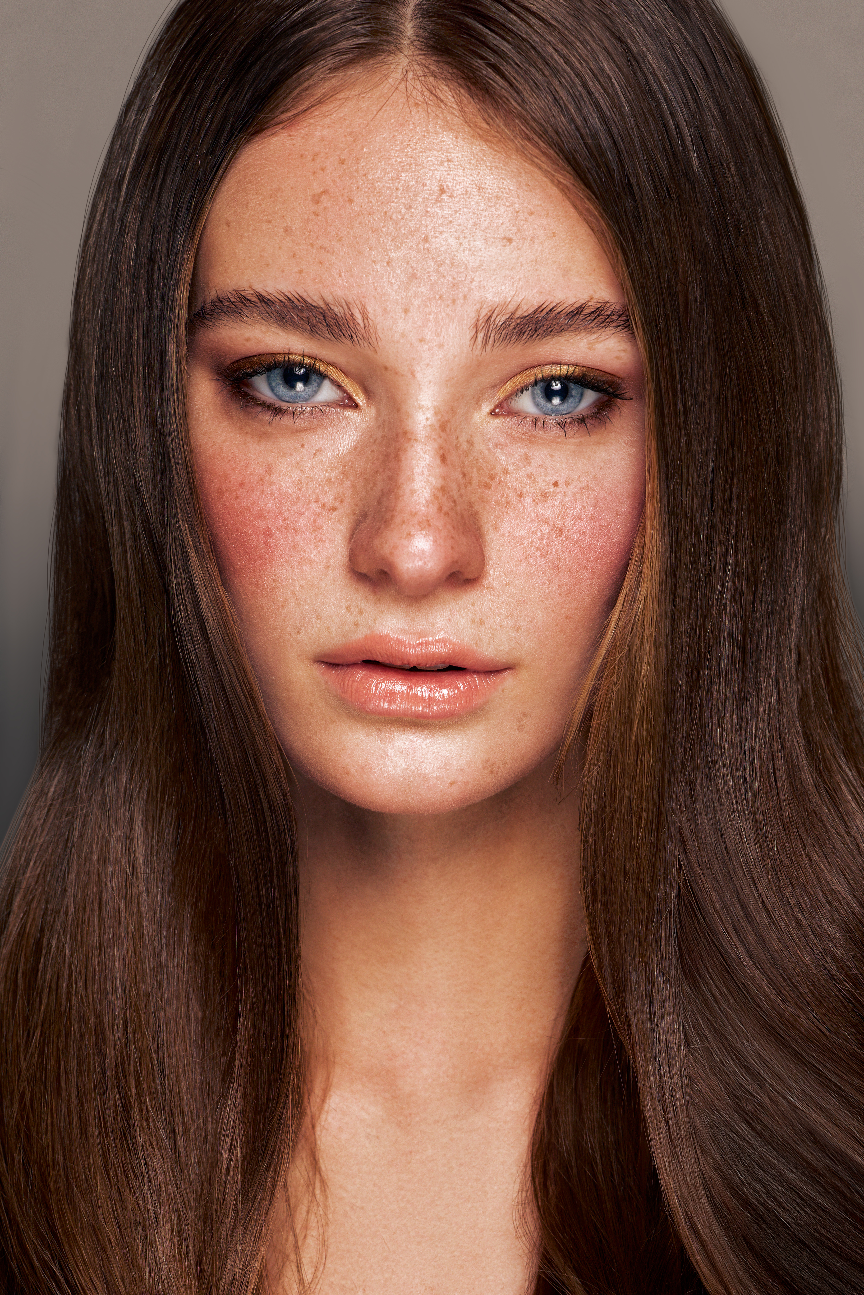 Close-up studio portrait of a young Caucasian woman with freckles and light blue eyes, showcasing beauty and makeup with long brown hair.