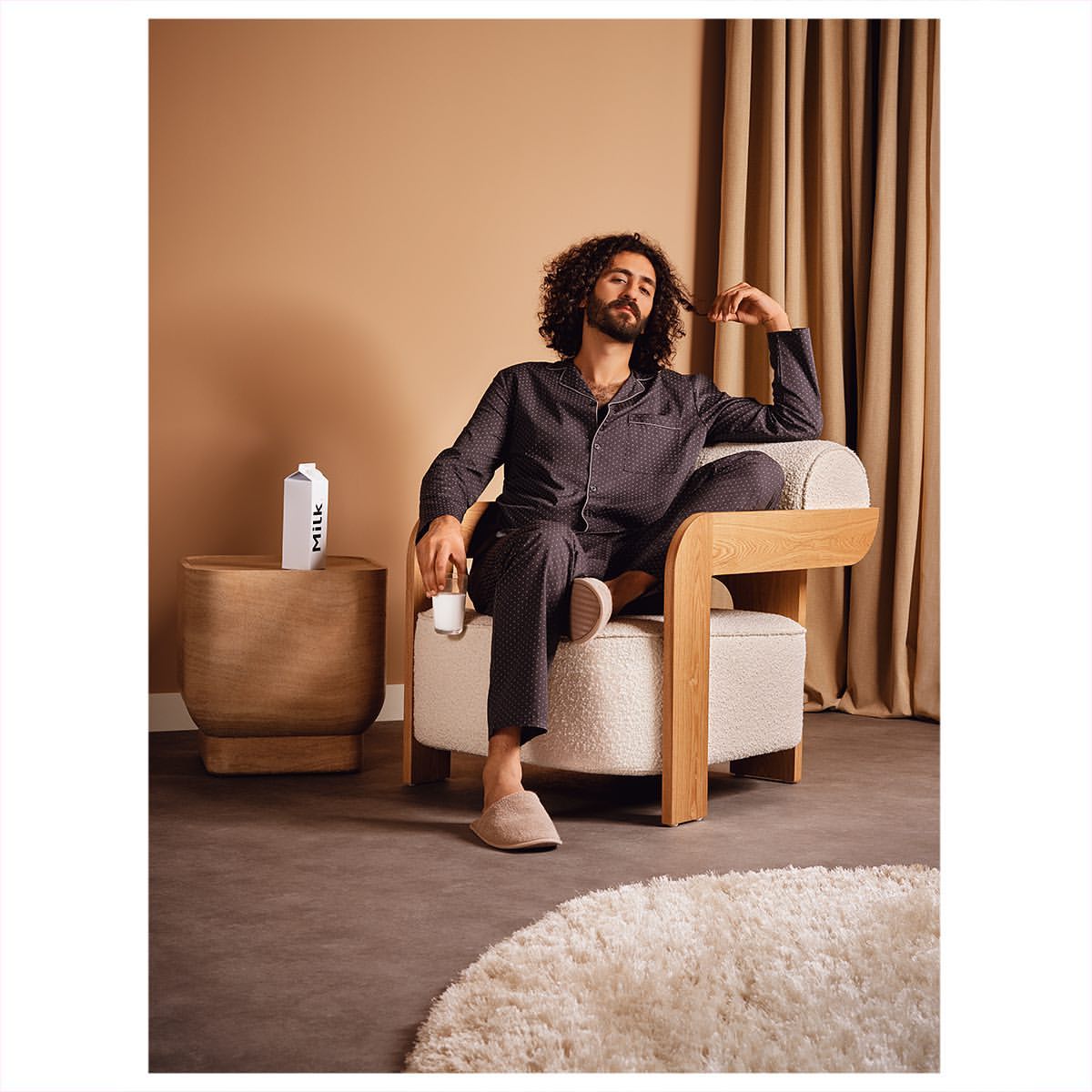 An adult male with curly hair relaxing in a modern chair while holding a small carton of milk in a neutral-toned room.