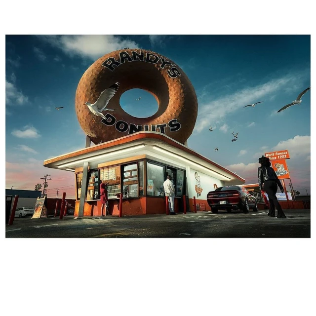 Randy's Donuts storefront with an oversized donut sign, people, and a car in the parking lot under a blue sky with scattered clouds.