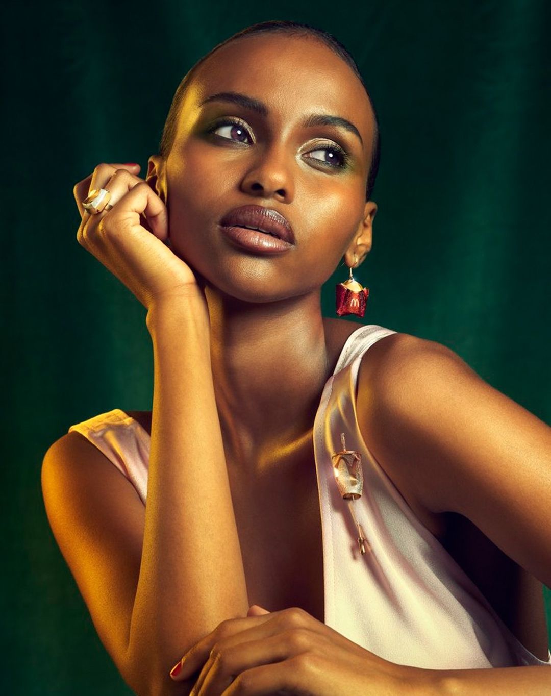 Close-up studio shot of a Black woman with bare shoulders wearing novelty jewelry against a green backdrop, gazing off camera.