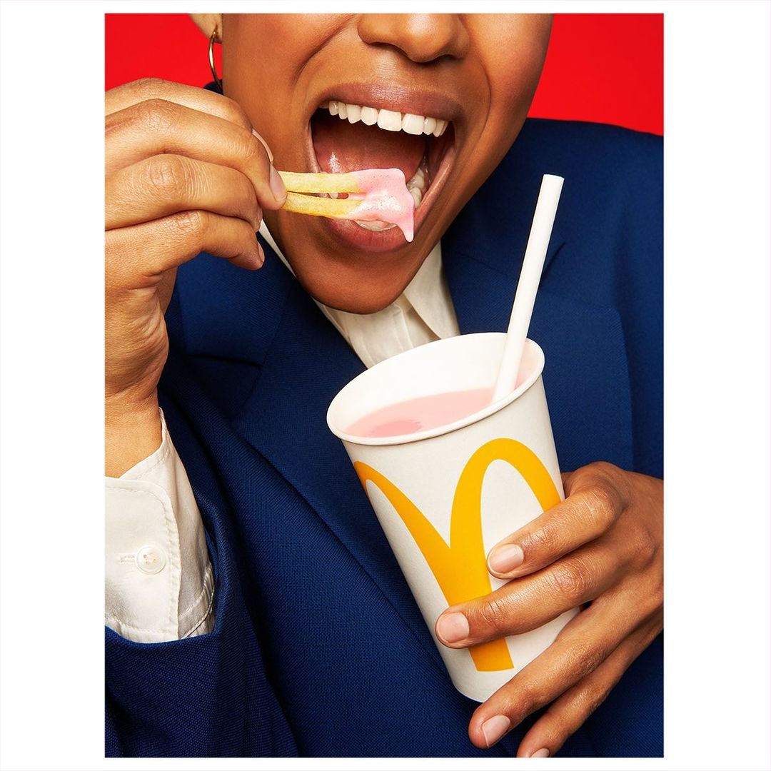 Person dips french fries in pink milkshake in a white paper cup with the McDonald's logo against a red background.