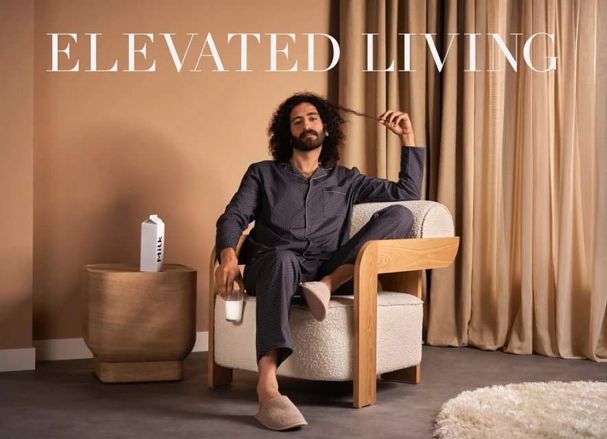 An adult male with curly hair lounges in pajamas, holding a glass of milk, in a light-filled interior setting with a carton of milk.